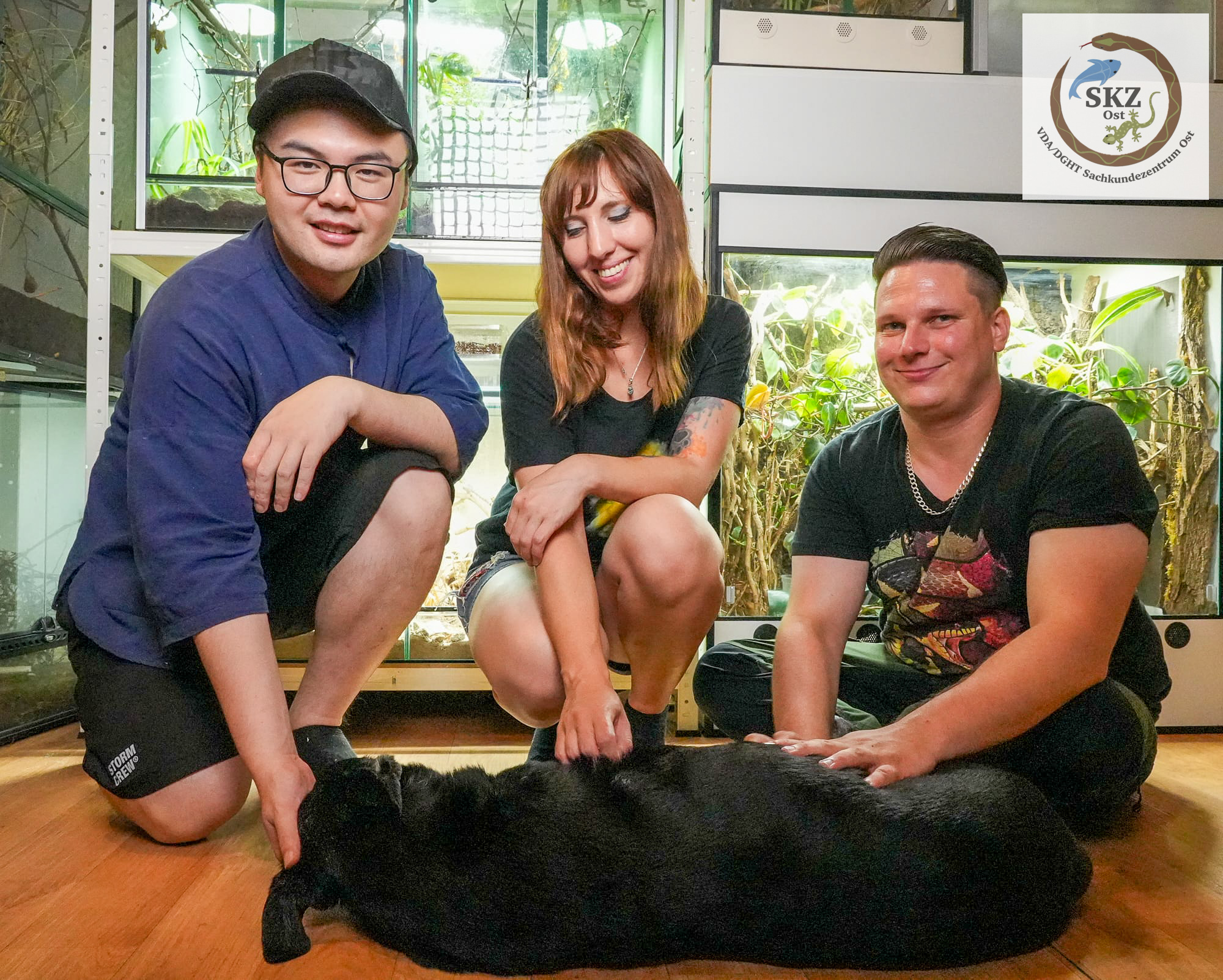 Drei Personen, eine Frau und zwei Männer, hocken vor mehreren Terrarien und streicheln einen Hund