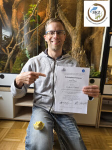 Ein erfolgreicher Teilnehmer der Schulung sitzt auf einem Stuhl vor einem großen Terrarium und zeigt sein Zertifikat