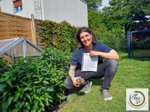 eine erfolgreiche Teilnehmerin unserer Schulung hockt im Garten und zeigt ihr Zertifikat und eine Schildkröte
